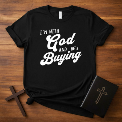 Black t-shirt with 'I'm with God and He's Buying' text on a wooden surface with crosses and a book.
